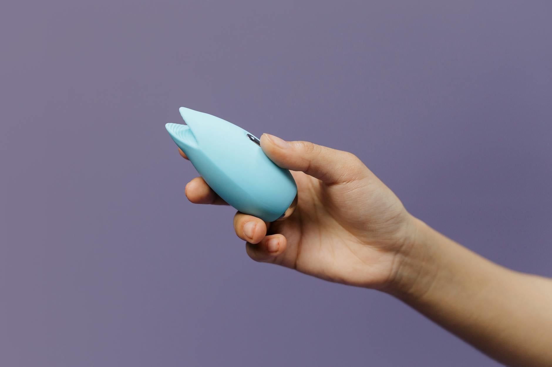 A blue silicone clitoral vibrator held in hand against a solid purple background, showcasing modern sexual wellness